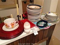 Photo showing red chargers, a cup and saucer with a cartoon-like design, white bowls with gold and blue trim stacked, and an egg-shaped shaker.