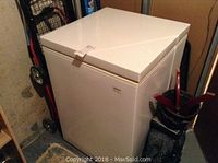 White Kenmore chest freezer with closed lid, situated in a basement or storage area next to a dolly and golf bag.