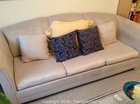 Front and side view of beige Bauhaus sofa bed with four decorative cushions on it placed in a carpeted room.