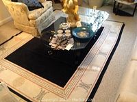 Photo showing the full area rug beneath a glass coffee table with a black center and beige border with symbols, highlighting general wear and the setting.