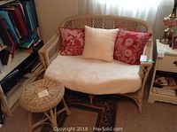 Wicker settee with cream seat cushion and three decorative pillows in red and beige tones, placed near window with sheer curtains.