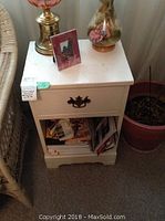 White painted night table with decorative drawer handle and open shelf below filled with books and papers, showing wear.