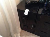Close-up of black metal filing cabinet skewed in a corner with visible drawers, lock, and key inserted.