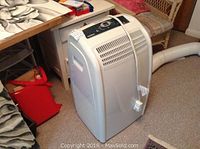 Full view of NOMA portable air conditioner standing on carpeted floor near furniture, plugged in with vent hose attached.