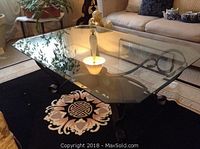 Image showing the beveled glass coffee table on a black decorative rug with light reflections on surface, metal legs visible underneath with curled ends.