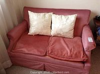 Red fabric loveseat with two white decorative pillows on top and visible wear on seat cushions, including an armrest cover on the right side.