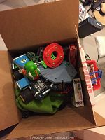 Photo of an open cardboard box filled with various toys including a green plush Muppet puppet and plastic toy parts such as a red circular piece and blue vehicle components.