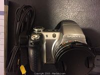 Front view of the silver and black Sony Cybershot DSC-H5 digital camera showing lens cover and part of the strap, with a coiled black cable.