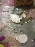 Overview of dining lot contents on floor showing soup tureen with lid and ladle, white water jug, clear glass serving tray, gold-toned metal serving tray, small clear dishes, and bubble-wrapped glassware.