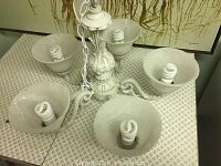 Complete view of the white ornate chandelier showing all five frosted glass light shades with spiral bulbs installed. The chandelier is placed on a patterned surface with a decorative painting in the background.