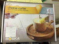 Box of Boska Holland cheese curler showing product image, brand name, and product usage with a wooden base and rotary curler tool.