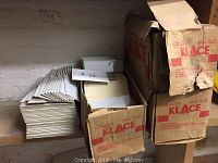 Boxes of grey ceramic wall tiles stacked on a shelf showing front and tops of boxes labeled Klace and loose tiles stacked beside them.
