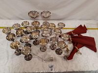 Overview of 22 silver plated stemmed glasses with 4 folded red cloth napkins in silver plated napkin rings alongside a long handled silver plated jigger.