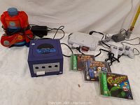 Photo of lot items: red and blue PSI Air Blaster toy with power adapter and remote, purple Nintendo GameCube console with attached controller and power adapter, white Sony PlayStation console with two controllers, and three PlayStation game cases including Tomb Raider III, Tomb Raider II, and It's Bad Frogger.