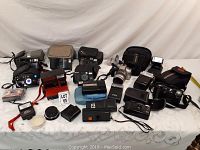 Overall lot of cameras, cases, accessories arranged on a table with white cloth background.