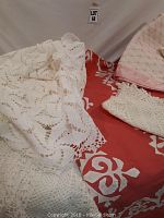 Various tablecloths and a blanket displayed, showing red white floral tablecloth, white lace tablecloths, pink patterned fabric, and beige embroidered cloth.