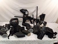 Overall view showing paintball gun with hopper, masks, holsters, and tactical gear on a white surface.