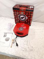 Red dog cookie maker with attached power cord, placed on a white surface alongside a metal storage tin with dog biscuit branding, four dog-shaped clear plastic cookie cutters, a wooden tool, and an instruction manual.