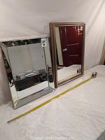 Two framed rectangular mirrors placed on a table, showing the front side. One mirror has a mirrored beveled edge frame, the other has a wooden frame.