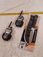 Photo showing two black and silver handheld walkie talkies and a packaged large survival knife with black handle and sheath.
