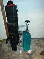 Black Wilson hard travel golf bag with broken lock, box and green golf bag hand cart behind.