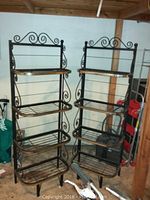 Two black metal bakers racks side by side against a wall in a storage room. Each has four metal bar shelves and decorative upper scroll designs.