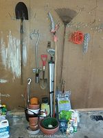 Photo showing 3 shovels of different sizes, a garden scraper, deck sprayer, several flower pots and gardening containers on the floor, and an orange extension cord and metal rake hung on the wall.