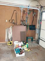 Wide shot showing entire collection of yard tools hanging on pegboard including pick axe, shovels, rake, and pest control products on floor