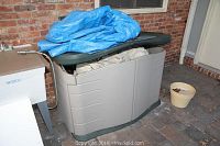 Closed resin deck box with blue tarp partially on top and beige plastic flower pot beside it containing soil. Shows storage setup outdoors against brick wall.