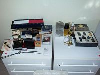 Wide view of the men's accessories including bolo tie, cuff links, button covers, travel belt, Perry Ellis tie case, and assortments of small fragrance bottles and boxes arranged on two white drawer tops.