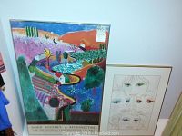 Framed colorful David Hockney poster with winding pathways and nature imagery alongside smaller framed signed artwork of stylized faces.