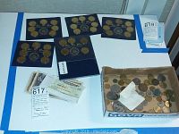 Wide shot of the various items including multiple blue cards with brass presidential coins, a box of mixed foreign coins, and several paper currencies.