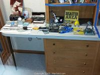 Wide view of white desk with mixed office supplies and miscellaneous items arranged on top and under bookshelf, including calculators, hole punches, booklet, wristbands, clock, and signs.