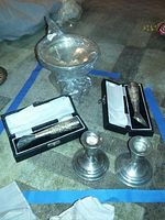 View of two weighted sterling silver candlesticks, sterling Fisher candy bowl in plastic wrap, and two boxed sterling silver bud vases on carpet.
