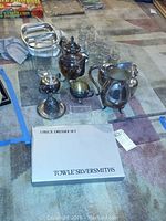 View of silverplate pitcher, teapot, sugar bowl, creamer, glass tumblers, and Towle Silversmiths 3-piece dresser set box arranged on carpet.