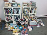 Wide shot of multiple white shelves filled with books and stacks of books scattered around the base.