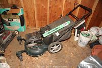 Full view of the gas powered lawn mower with grass collection bag attached and mulcher blade included, positioned in a shed or garage on dirt floor.