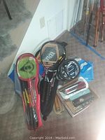 Full view of sports equipment grouped showing badminton set in red bag, tennis racquets with protective bags, bike helmet, and golf accessories.