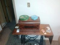 Antique wooden cabinet housing an older sewing machine, sitting on wrought iron treadle base with embossed 'HOUSEHOLD' on both sides. Cabinet lid closed.