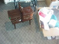 Wooden floor-standing tiered sewing box with handle and metal hinges on sides, showing surface wear and dings, set on carpeted floor near cardboard box with fabric and sewing supplies.