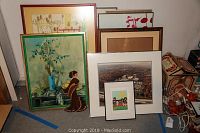 Photo of eight framed prints and photos leaning against a wall, ranging in size. Includes framed images of Asian woman, local aerial views, and others.