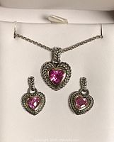 Close-up image showing heart-shaped sterling silver necklace pendant and matching earrings with magenta pink heart-shaped gemstones and gold outlines on a white cushioned background.