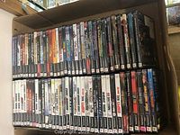 Photo showing over 60 PlayStation 2 games in their original cases, arranged in two rows inside a cardboard box, with visible cover art and spine labels.