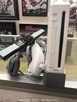 Photo showing white Nintendo Wii console upright with white Wii Remote joystick wrapped with cable and black sensor bar beside it.