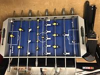 Top-down photo of foosball table showing blue playing field, plastic players in white and yellow on metal rods, gray handles, and black detached legs.