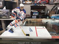 Photo of mounted Wayne Gretzky figurine in Edmonton Oilers uniform, displayed on a glass table in a room with various other items visible.