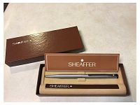 Photo showing the vintage silver-tone Sheaffer White Dot refillable ballpoint pen with blue ink cartridge inside the original burgundy box. The beige velvet interior holds the pen and the original Sheaffer pamphlet is displayed at the back of the case.