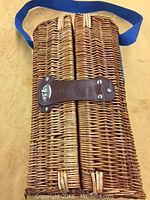 Full view of the wicker carrying case showing natural wicker weave, brown leather handle, and attached blue shoulder strap.