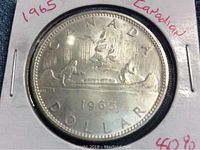 Close-up front (reverse) image of the 1965 Canadian silver dollar coin showing two voyageurs paddling a canoe with maple leaf above and date below.
