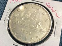 Close-up photo of the 1963 Canadian silver dollar coin showing voyageur canoe design and date '1963'. Coin protected in holder with handwritten notes.